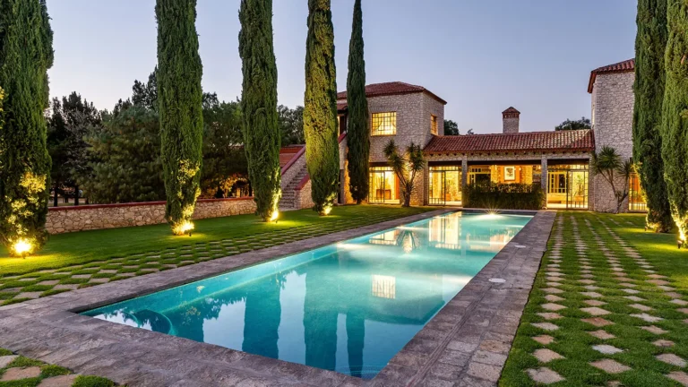 Rancho Las Sabinas, a luxury estate in San Miguel de Allende featuring a stone façade, landscaped gardens, and a long illuminated swimming pool at dusk.