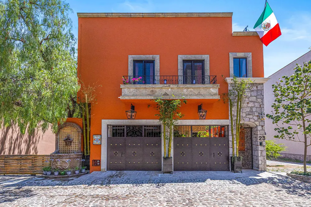 Two-story orange house with gray stone trim, a wrought-iron balcony, and a large dark gate on a cobblestone street; a Mexican flag waves from the roof.