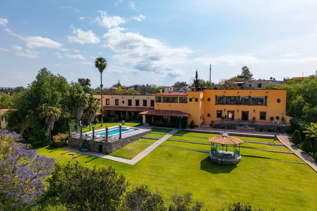 Mediterranean-style yellow villa with a rectangular blue pool, manicured lawn, palm trees, and a stone terrace in a sunny courtyard.